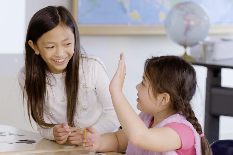 Two girls sit at a desk in a classroom, smiling and talking, with a globe and map in the background.