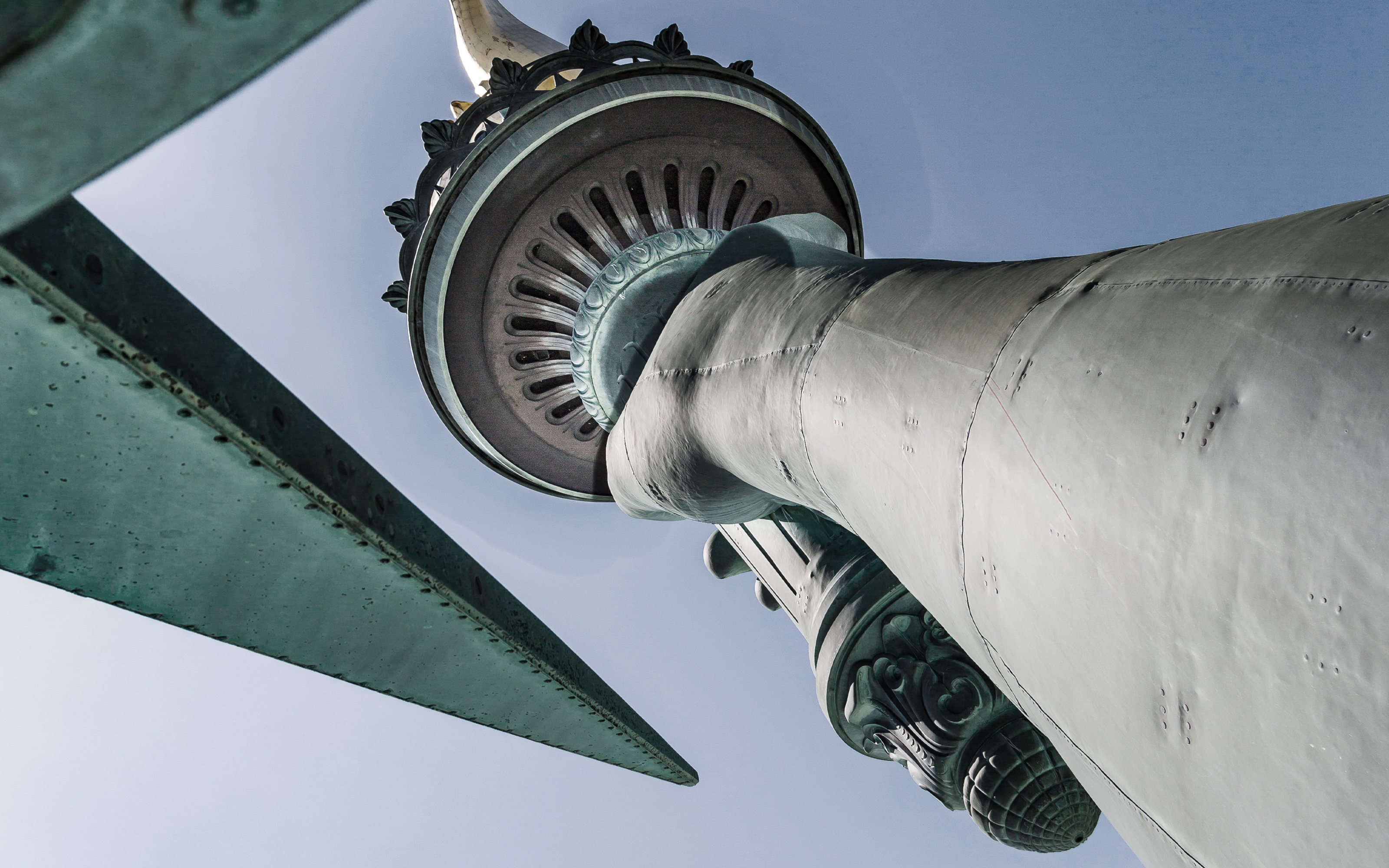 Close-up upward view of the Statue of Libertys arm, torch, and crown against a clear sky.