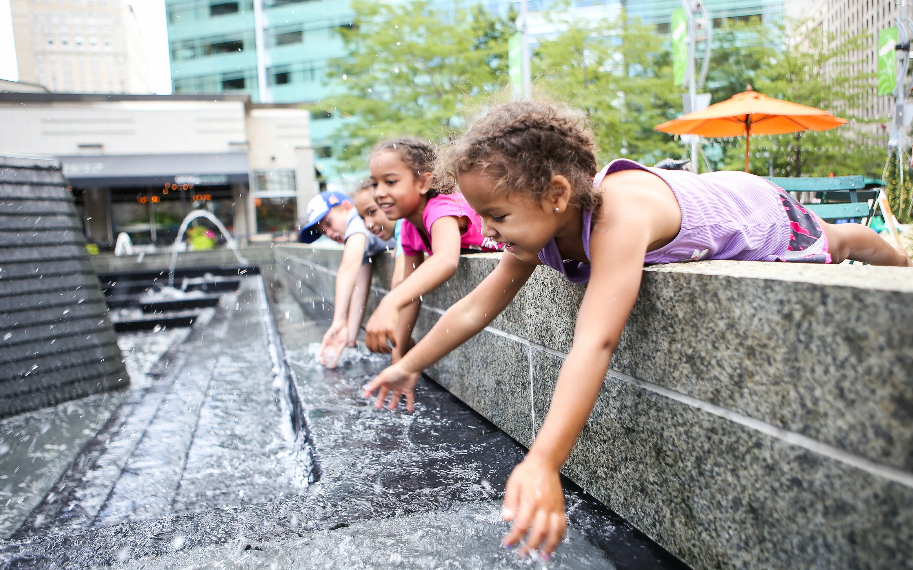 Three children lean over a stone ledge, reaching their hands into a playful urban fountain on a sunny day.