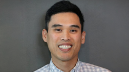 A smiling man wearing a light blue and white checkered shirt.
