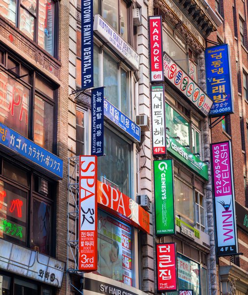 Colorful signs in English and Korean hang from buildings in a busy city street, advertising spas, karaoke, and salons.