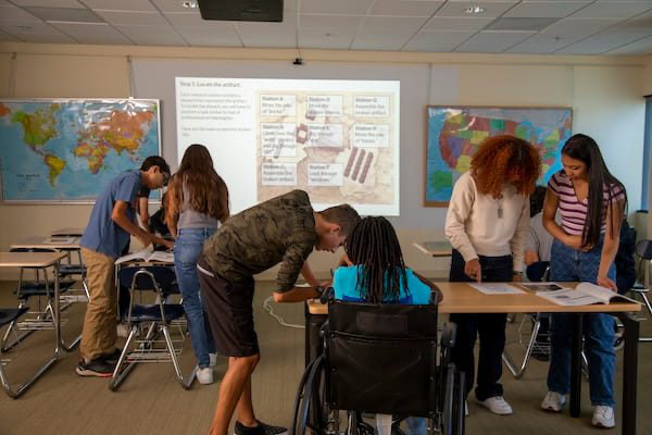 Six students, including one in a wheelchair, work together on assignments in a classroom with world maps on the walls.