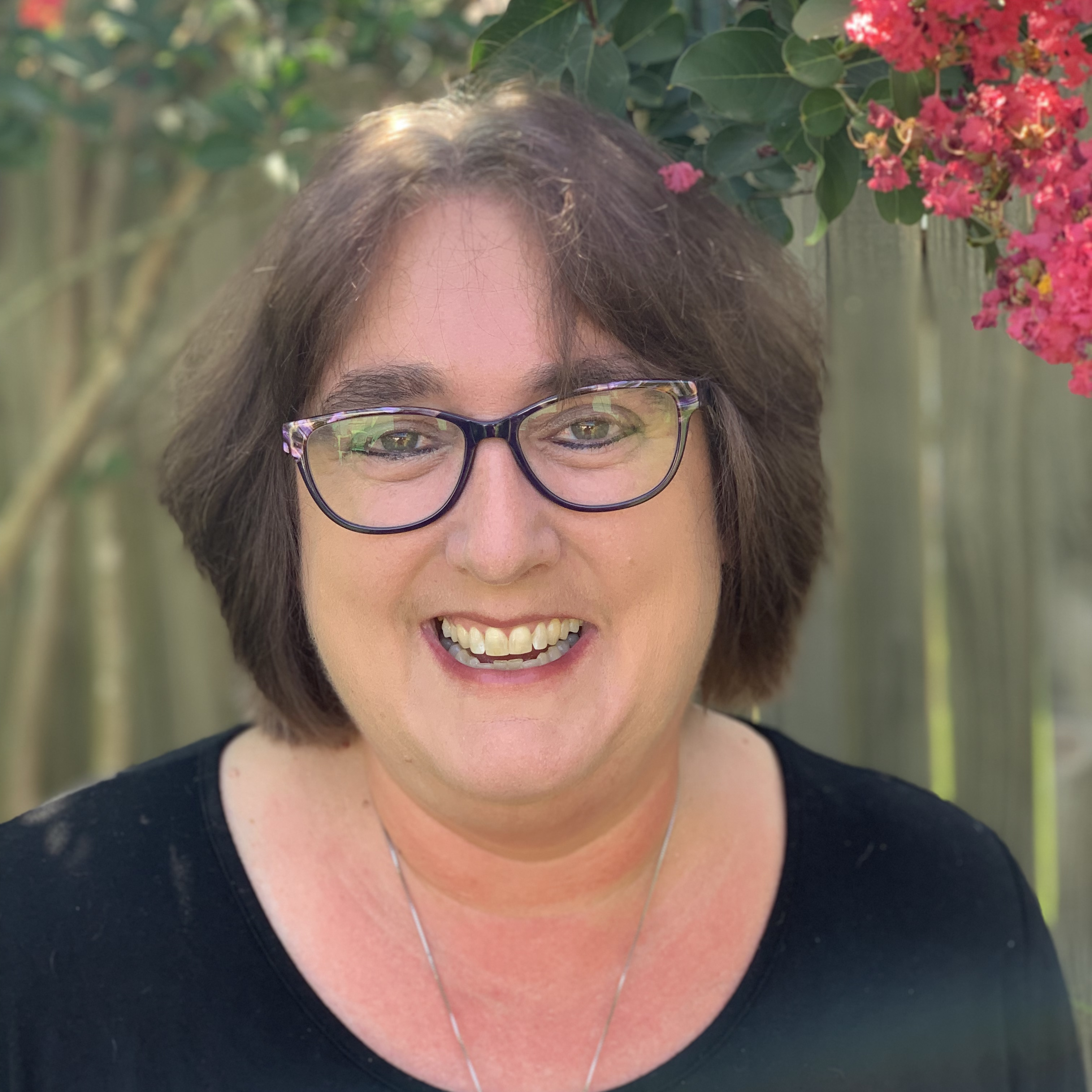 Smiling woman with glasses and short brown hair, wearing a black top, standing outdoors in front of a wooden fence.