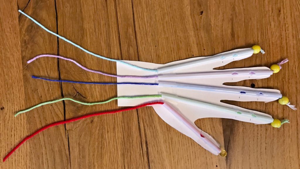 A paper hand with straws for fingers, each with yarn tendons and beads, lies on a wooden surface.