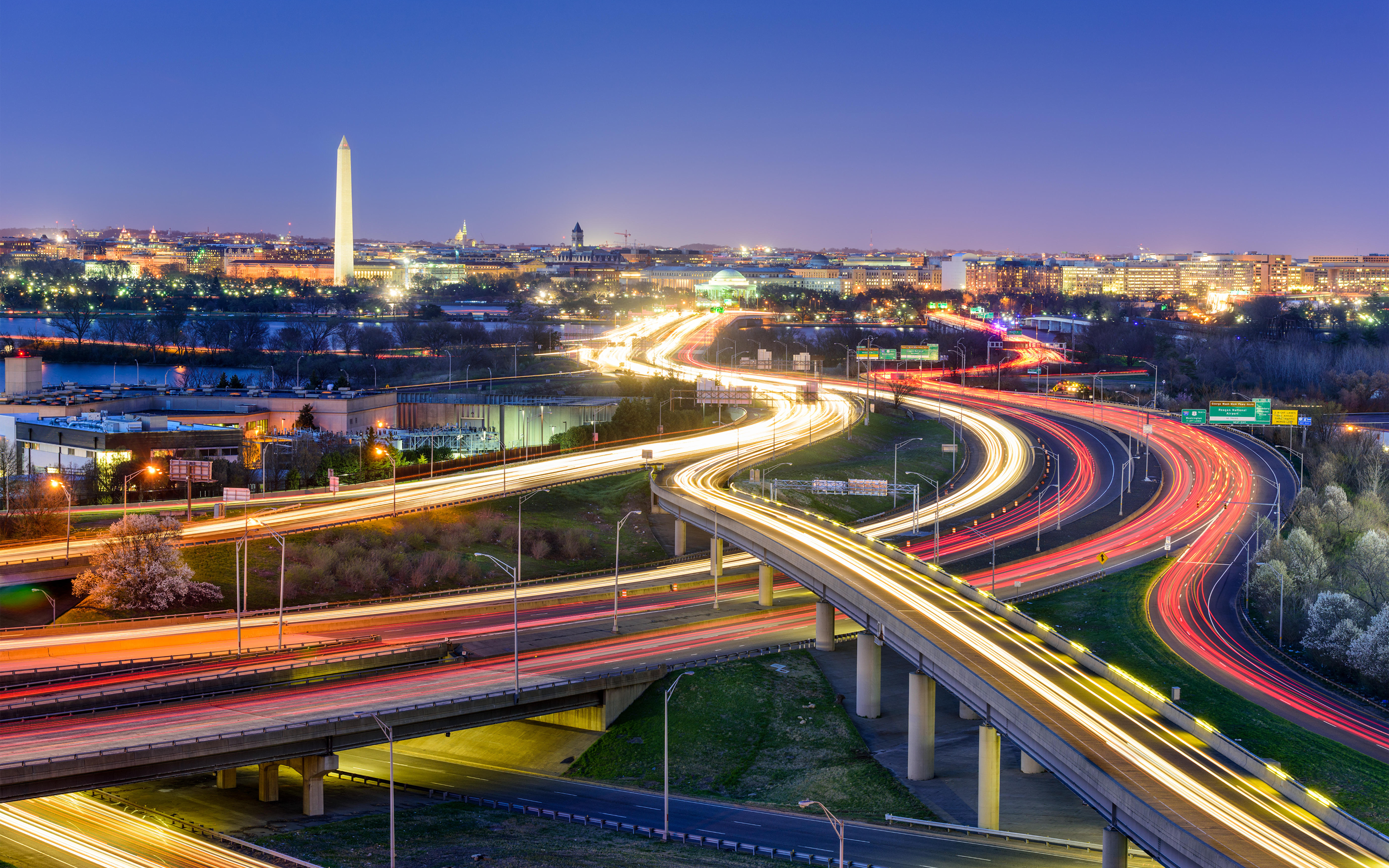 Cityscape of Washington, D.C. at dusk with light trails on highways and the Washington Monument in the background.