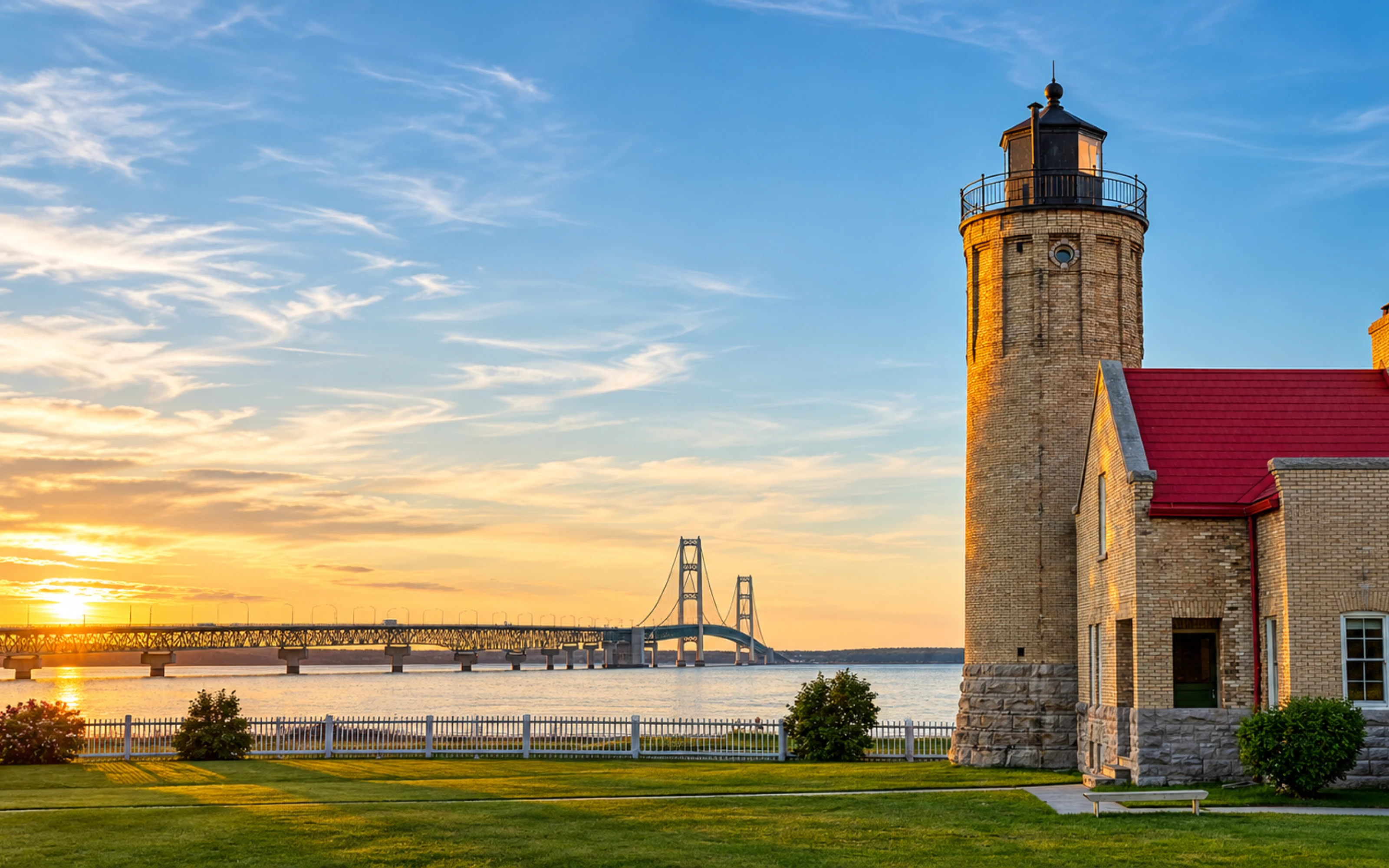 Brick lighthouse with red roof near a large bridge over water at sunset, with a grassy lawn in front.