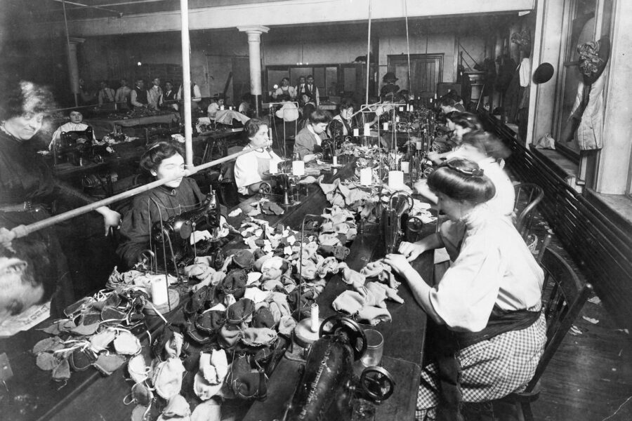 Women work at sewing machines in a crowded garment factory, surrounded by fabric and materials, early 1900s.