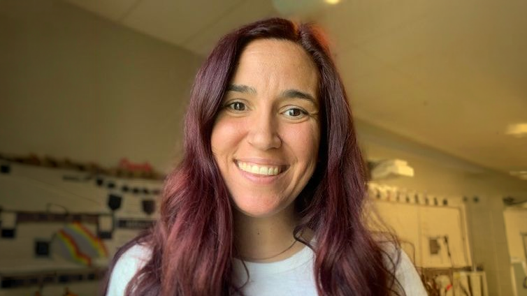 Woman with long wavy hair smiling indoors, classroom decor visible in the background.