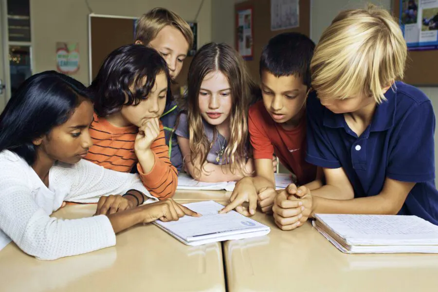 Six children sit at a desk, looking at and discussing something in an open notebook together.
