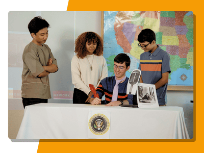 Four high school students gather around a table set up like a presidential signing ceremony, with a seated student holding a large pen near a microphone and a presidential seal on the table skirt, a US map on the wall behind them.