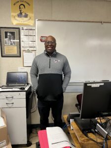Teacher Michael Washington stands in front of a white board and next to a file cabinet and desk